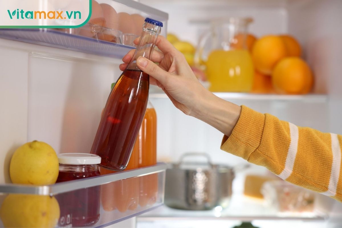 Đôi tay đặt bình thủy tinh có nắp chứa nước linh chi màu nâu hổ phách vào ngăn mát tủ lạnh sạch để bảo quản phần nước chưa uống.
