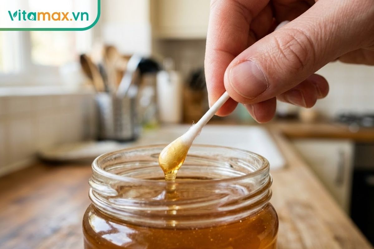 Cận cảnh bàn tay sạch đang cầm tăm bông y tế lấy một lượng nhỏ mật ong từ trong hũ thủy tinh, không để tay chạm vào mật ong.