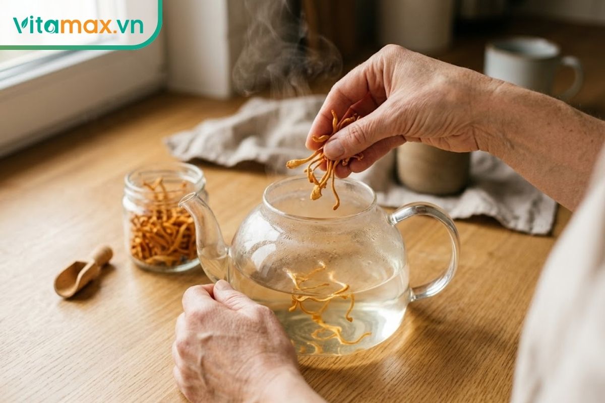 Đôi bàn tay đang cẩn thận thả những sợi đông trùng hạ thảo khô vào một ấm trà thủy tinh trong suốt chứa nước nóng trên bàn bếp.