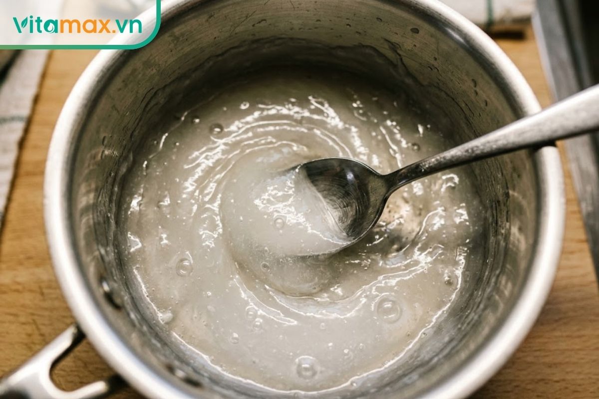 Cận cảnh một chiếc nồi nhỏ trên bếp, bên trong là hỗn hợp bột sắn dây đã được nấu chín, có màu trắng trong suốt, đặc sánh, đang được khuấy bằng một chiếc thìa.