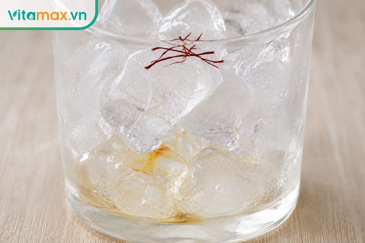 Cận cảnh viên đá lạnh tinh khiết đang tan từ từ bên trong một chiếc cốc thủy tinh nhỏ, đè lên những sợi nhụy hoa nghệ tây saffron màu đỏ sẫm đang bắt đầu tiết ra màu vàng.
