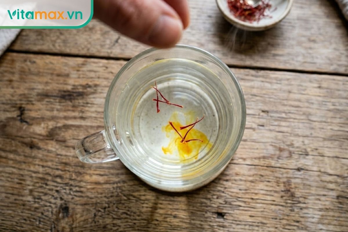 Góc nhìn từ trên xuống của một bàn tay đang nhẹ nhàng thả 4 sợi nhụy hoa nghệ tây màu đỏ thẫm vào một ly nước ấm, bắt đầu nhả màu vàng nhẹ.
