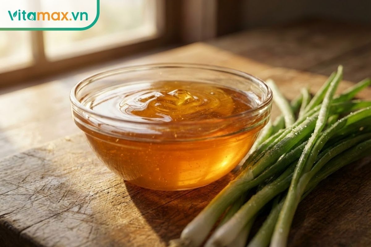 Chén thủy tinh đựng mật ong vàng sóng sánh đặt cạnh bó hành lá tươi xanh còn đọng nước trên bàn gỗ sồi.
