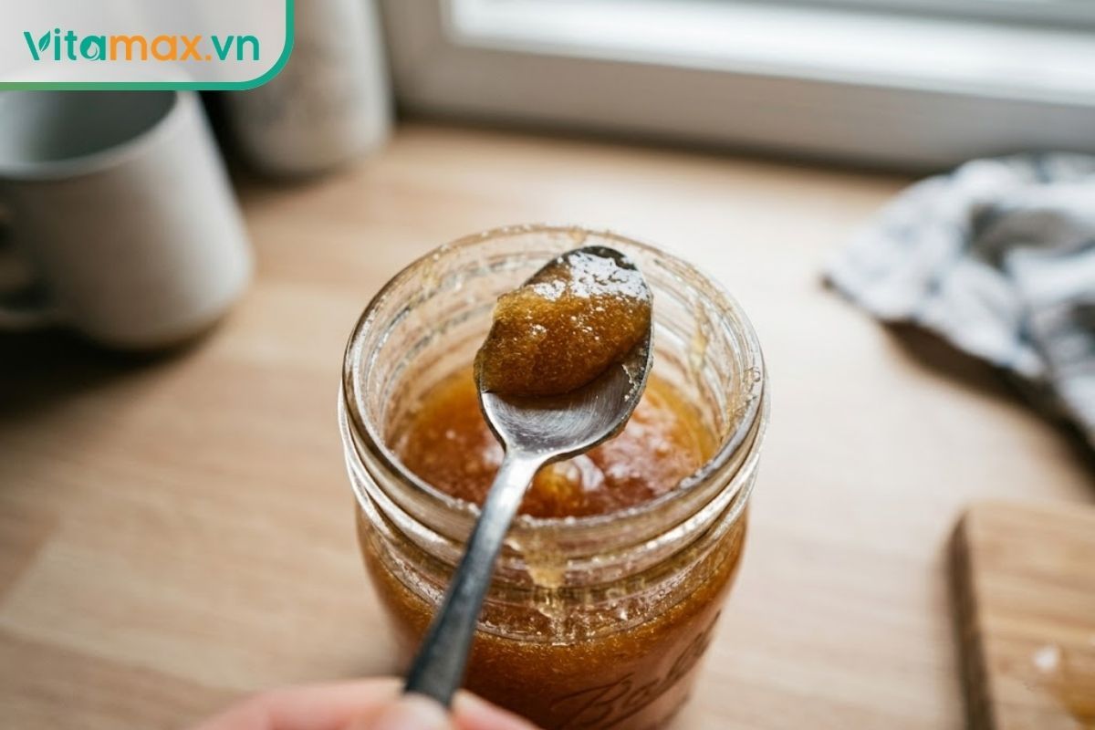 Cận cảnh một chiếc thìa cà phê bằng kim loại đang múc một lượng nhỏ mật ong vừa phải từ trong hũ thủy tinh ra ngoài.