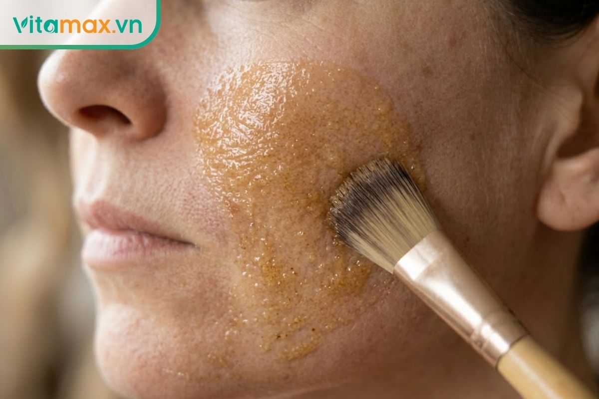 Cận cảnh một chiếc cọ chuyên dụng đang quét một lớp mỏng hỗn hợp mặt nạ nhân sâm và mật ong lên vùng má của một người phụ nữ, hỗn hợp màu vàng ngà bám đều trên da.
