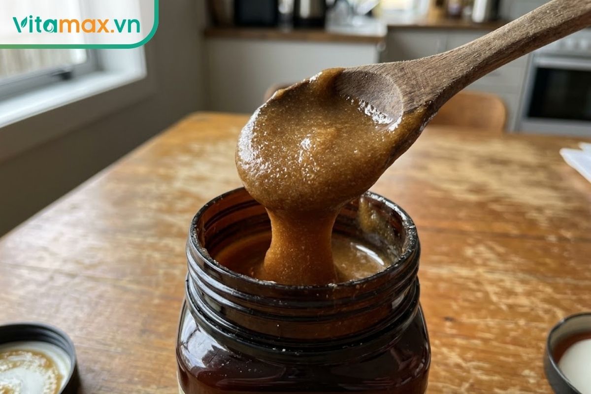 Cận cảnh một chiếc thìa nhỏ đang múc lớp mật ong Manuka đặc sánh, màu sậm đục từ trong hũ thủy tinh để dùng trực tiếp chữa ho