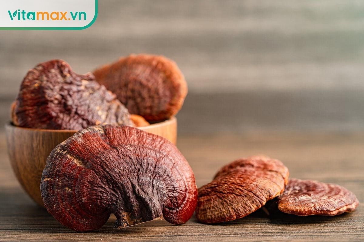 Nấm linh chi đỏ nguyên tai và nấm thái lát đặt trên bàn gỗ sáng, thể hiện màu nâu đỏ, hình dáng tai nấm và kết cấu tự nhiên.