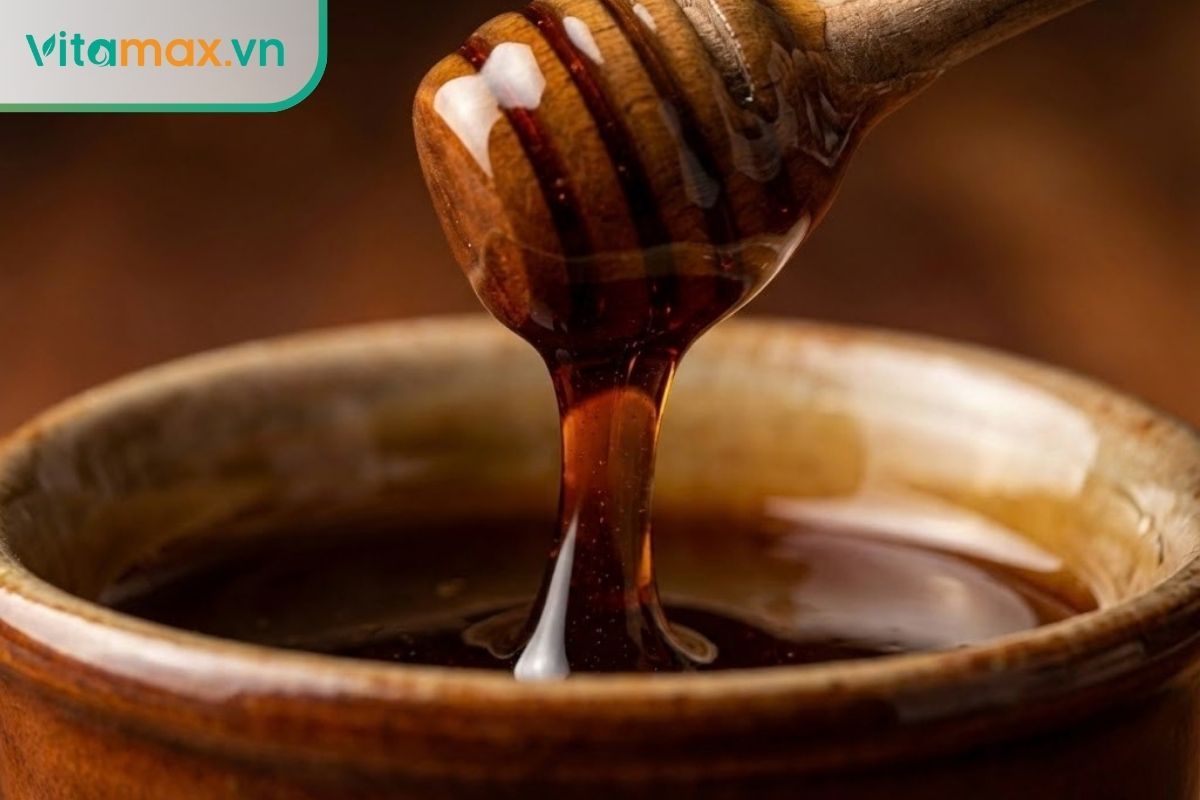 Cận cảnh dòng mật ong màu nâu đen chảy từ thìa gỗ xuống, chất mật sánh mịn, không tách lớp.