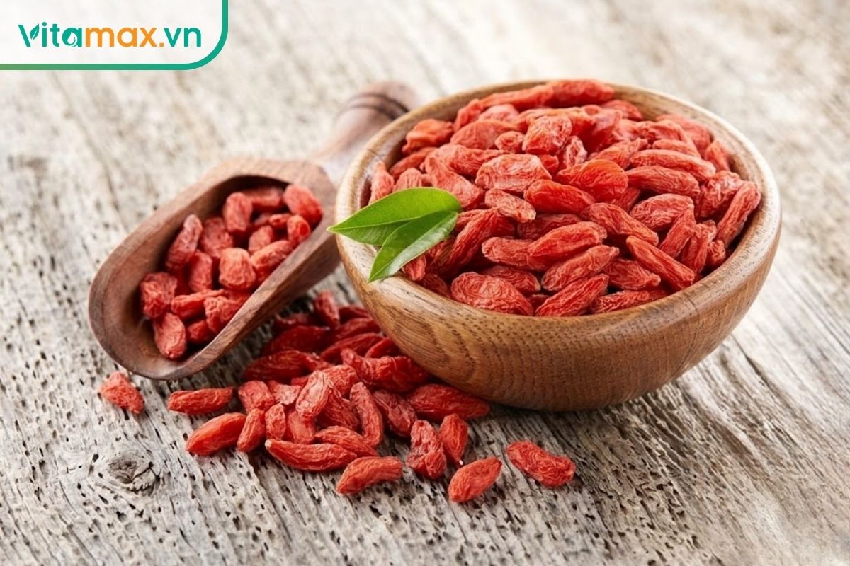 Kỷ tử khô màu đỏ cam được đựng đầy trong bát gỗ cùng thìa gỗ, nổi bật trên mặt bàn gỗ sáng tự nhiên.