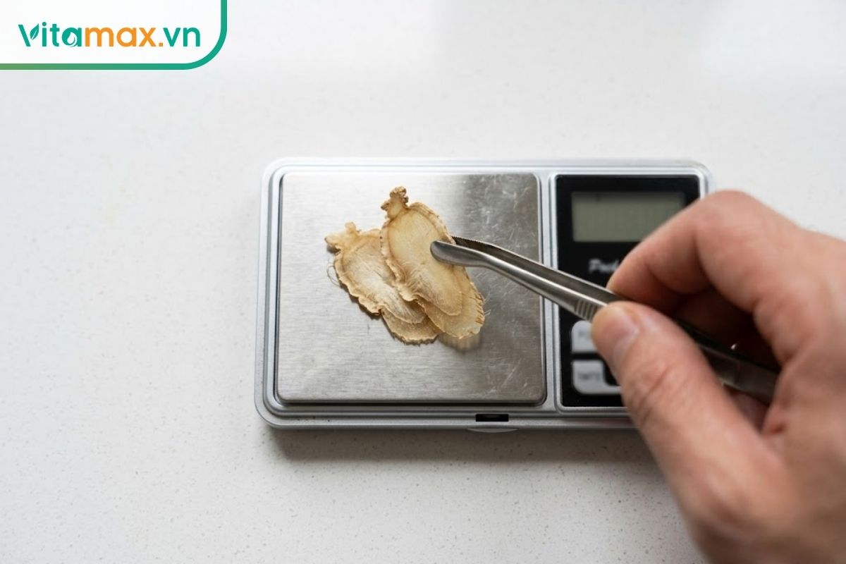 Cận cảnh một người đang dùng chiếc kẹp kim loại gắp hai lát sâm Ngọc Linh tươi rất mỏng đặt lên một chiếc cân điện tử mini nhà bếp màu bạc.