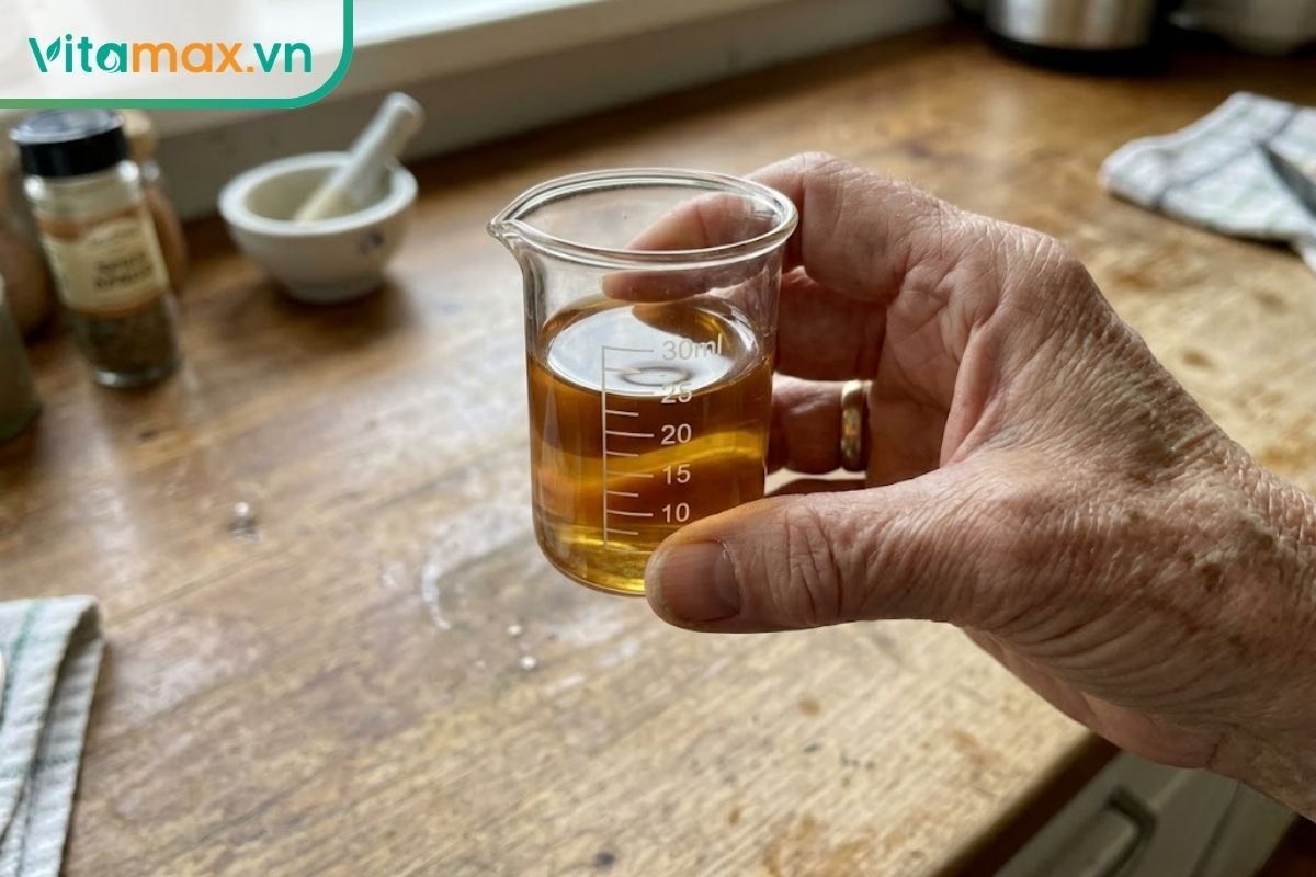 Cận cảnh tay người lớn tuổi cầm ly đong dung tích nhỏ chứa khoảng 20-30ml rượu sâm Ngọc Linh, hướng dẫn liều lượng sử dụng an toàn.