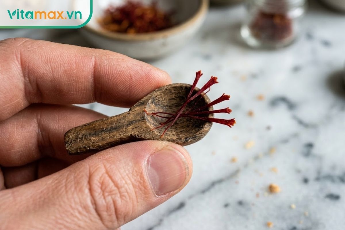 Cận cảnh một chiếc thìa gỗ nhỏ đang múc chính xác 5 sợi nhụy hoa nghệ tây màu đỏ thẫm trên nền bàn bếp sáng màu, thể hiện liều lượng an toàn.