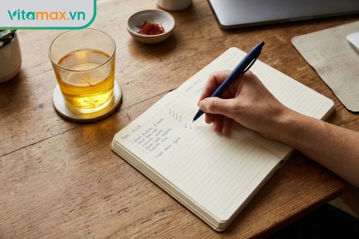 Góc chụp từ trên xuống mặt bàn gỗ, một tay đang cầm bút ghi chép vào cuốn sổ tay nhỏ bên cạnh một tách trà saffron màu vàng nhạt.