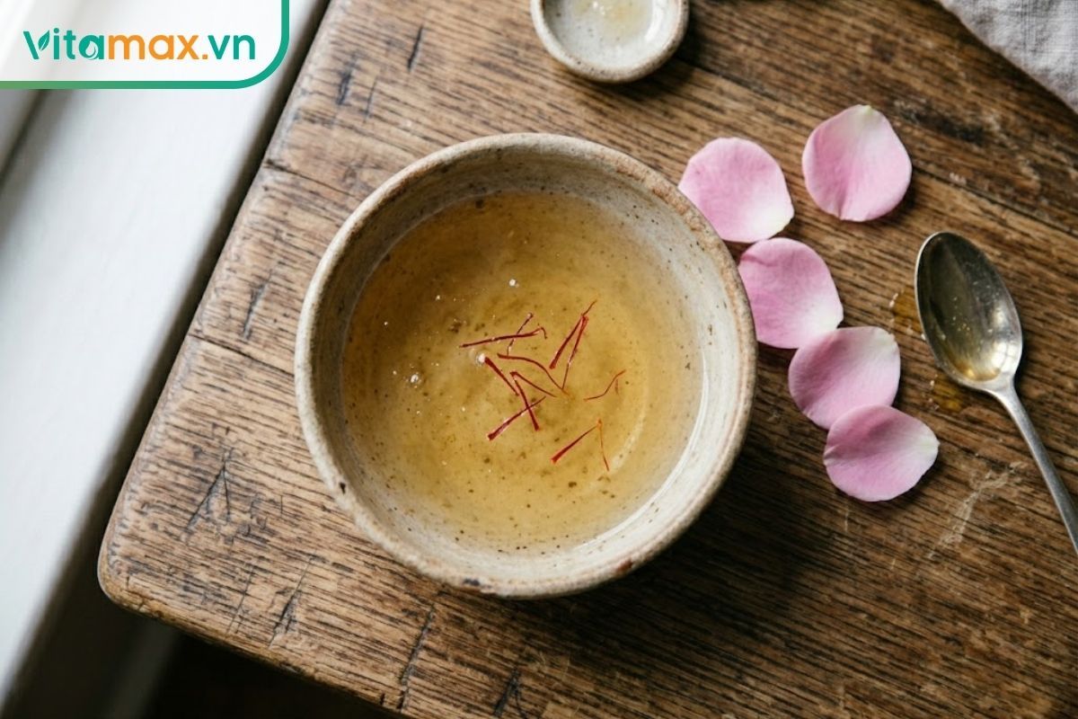 Chén sứ nhỏ chứa hỗn hợp mặt nạ nhụy hoa nghệ tây, mật ong và nước hoa hồng lỏng nhẹ, trong vắt, màu vàng ươm phù hợp cho da nhạy cảm.
