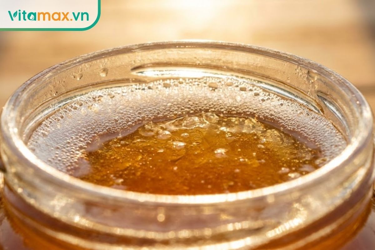 Cận cảnh lớp bọt khí trắng mịn nổi trên bề mặt hũ mật ong nguyên chất màu vàng hổ phách.