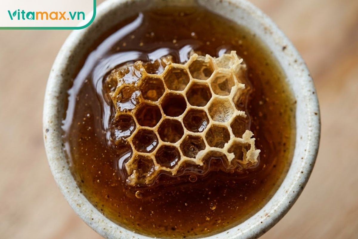 Cận cảnh một thìa mật ong sẫm màu đặc sánh có lẫn một miếng sáp ong tự nhiên nhỏ, đặt trên mặt bàn gỗ.