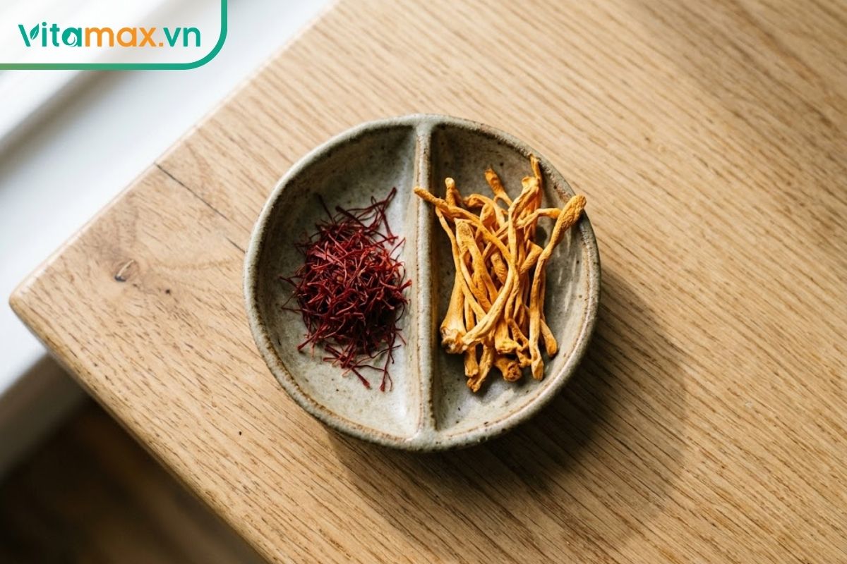 Đĩa gốm nhỏ chia làm hai nửa chứa sợi nhụy hoa nghệ tây saffron đỏ thẫm và nấm đông trùng hạ thảo màu vàng cam, đặt trên mặt bàn gỗ sáng màu.