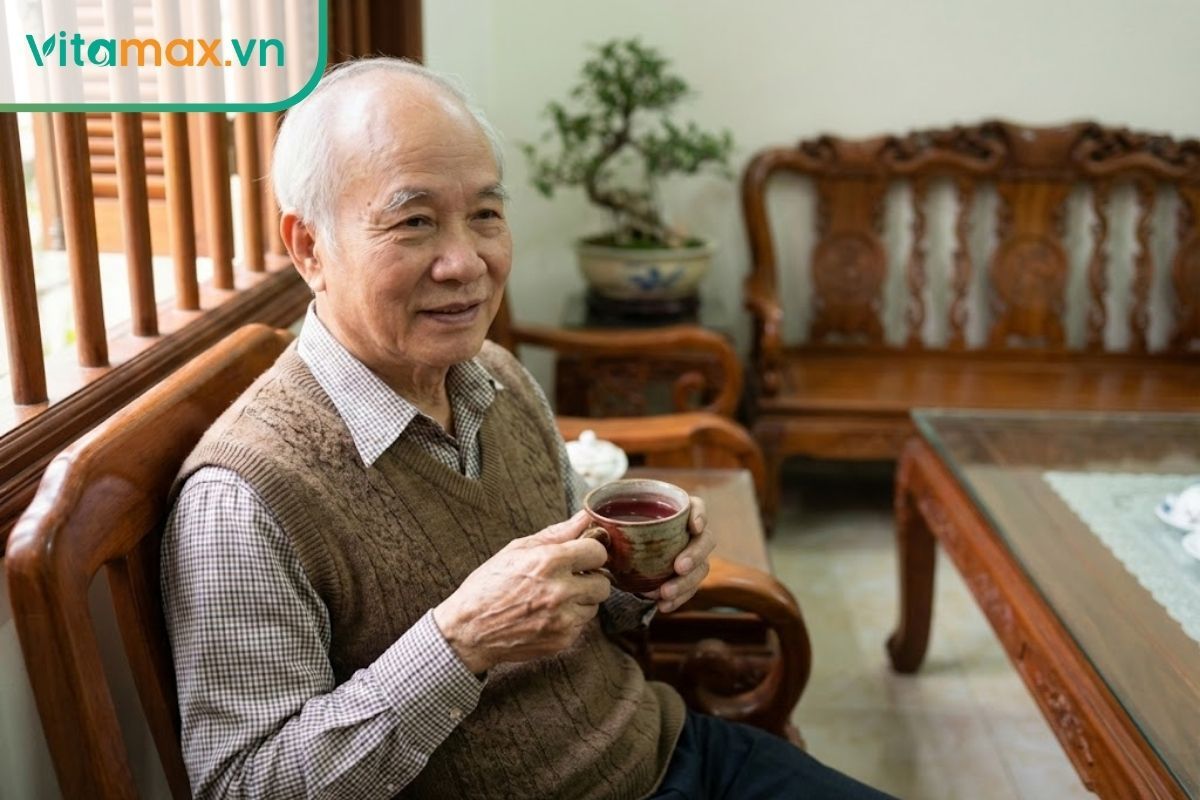 Cụ ông người Việt Nam tóc bạc phơ, mặc áo len gile ấm áp đang cầm cốc trà hồng sâm nóng, ngồi thư thái tại phòng khách.