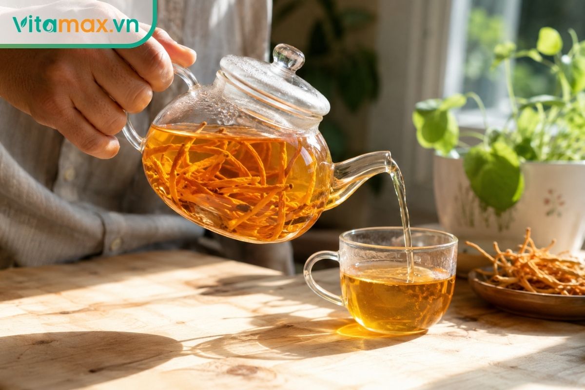 Tay một người đang rót nước trà màu cam óng ánh từ bình thủy tinh có chứa nhiều sợi đông trùng hạ thảo ra một chiếc chén nhỏ, ánh nắng ban mai chiếu rọi rực rỡ.