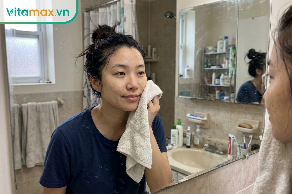 Hình ảnh đời thường một cô gái đang dùng khăn bông thấm nhẹ lên da mặt ướt trước gương, không chà xát mạnh để tránh tổn thương da.