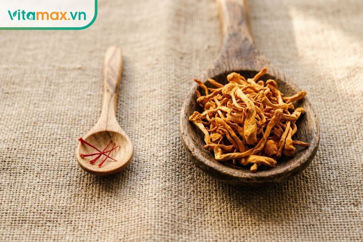 Cận cảnh một chiếc thìa gỗ nhỏ xíu chứa 3 sợi saffron đỏ cạnh một chiếc thìa gỗ lớn hơn chứa một nhúm nấm đông trùng hạ thảo vàng cam trên nền vải lanh.
