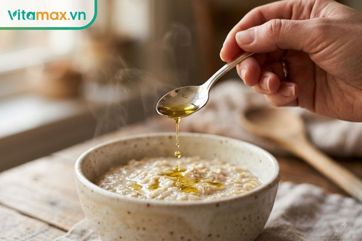 Cận cảnh một chiếc thìa nhỏ đang nhỏ vài giọt dầu ăn dinh dưỡng màu vàng óng vào một bát cháo nóng hổi vừa nấu xong cho trẻ.