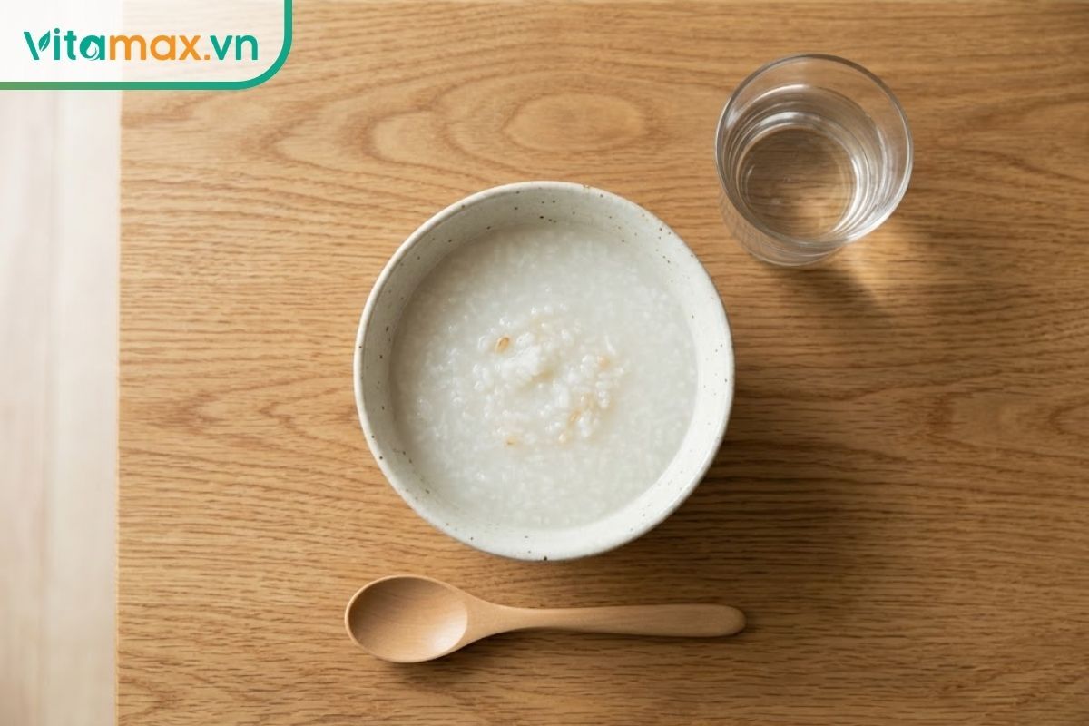 Một bát cháo yến mạch hoặc súp mềm mịn đặt trên bàn ăn bằng gỗ, bên cạnh là một cốc nước lọc, tông màu ảnh ấm áp và mang cảm giác chữa lành.