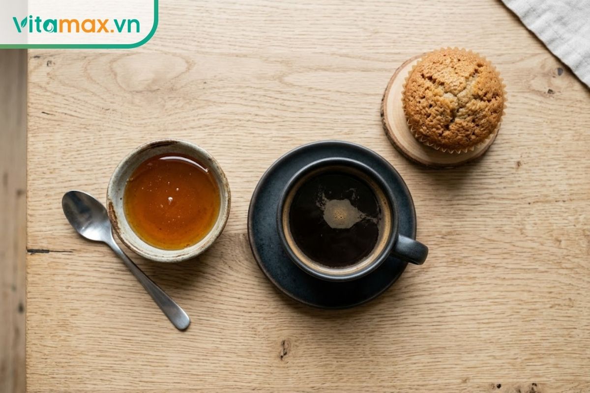 Một chiếc bát nhỏ đựng mật ong đặt cạnh một tách cà phê đen và một chiếc bánh ngọt trên bàn ăn sáng, thể hiện các nguồn cung cấp đường khác nhau.