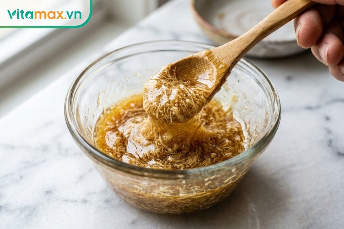 Hỗn hợp sâm tươi xay nhuyễn màu vàng ngà đang được khuấy đều cùng mật ong đặc sánh trong một chiếc bát thủy tinh nhỏ bằng muỗng gỗ.